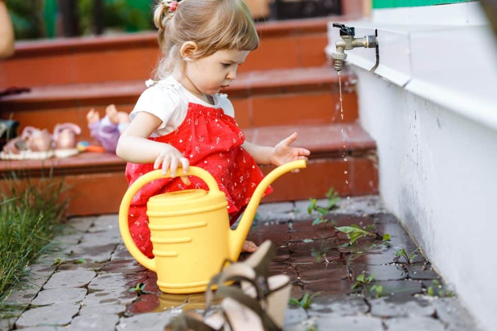 A little girl dressed in a sundress is drawing water in a watering can in the garden next to the